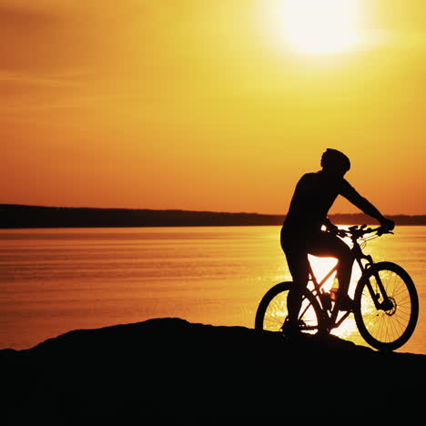professional male biker gets off the bike near the river at a beautiful sunset. silhouette of a sporty cyclist in helmet on the evening water background at orange setting sun.