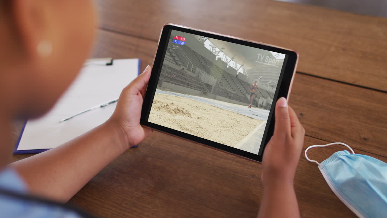 태블릿에서 육상 하이 점프 이벤트를 시청하는 얼굴 마스크와 함께 테이블에 앉아있는 여성의 복합