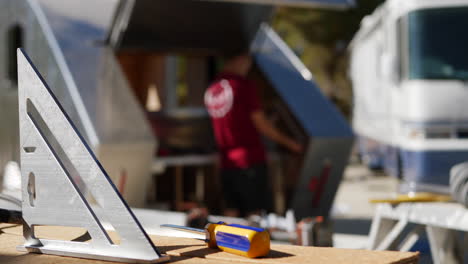 cierre de un taladro inalámbrico con una broca al sol mientras un trabajador de la construcción construye un remolque de viaje personalizado en un lugar de trabajo en el fondo con espacio de copia
