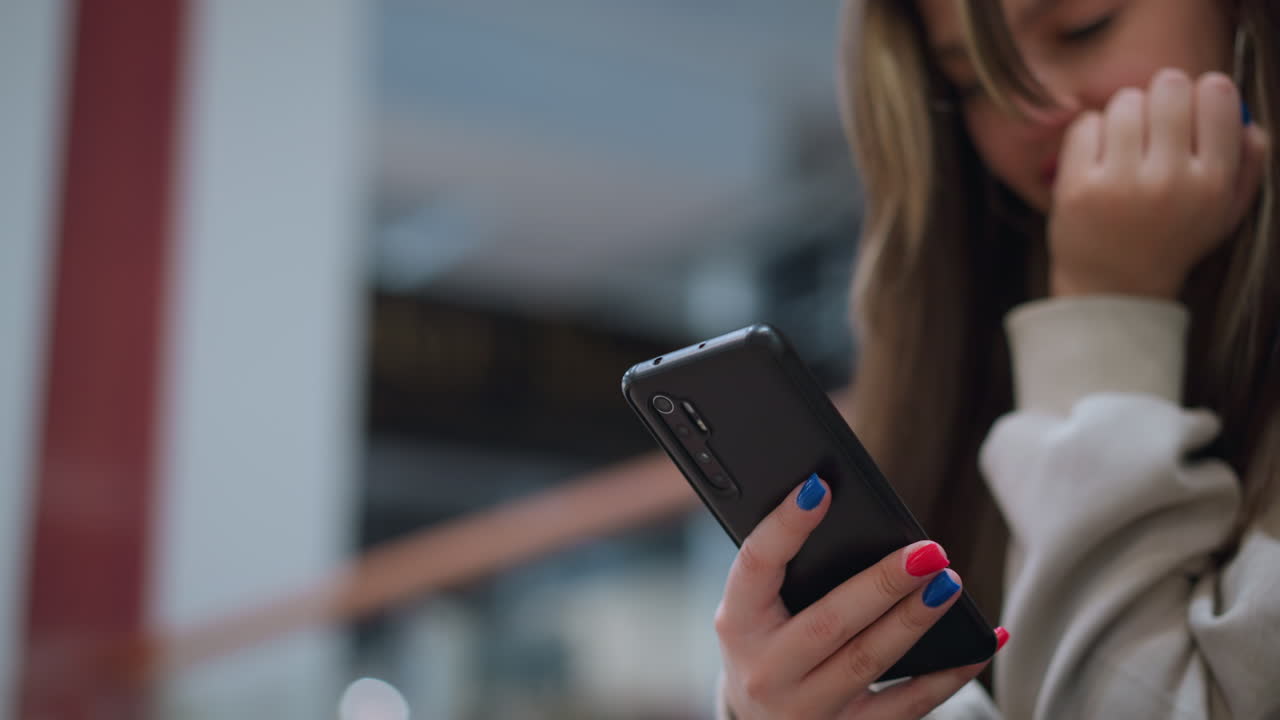 Young lady with colorful nail paint operating phone indoors resting chin on hand showing focus and engagement with digital communication reflecting youth culture