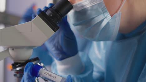 female lab worker wearing ppe analysing samples with microscope holding test tube labelled covid-19