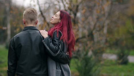 joven pareja romántica caminando en el parque de otoño durante el día. vista trasera de un joven hombre rubio en chaqueta de cuero abrazando a su novia. pareja amorosa hablando y pasando tiempo juntos