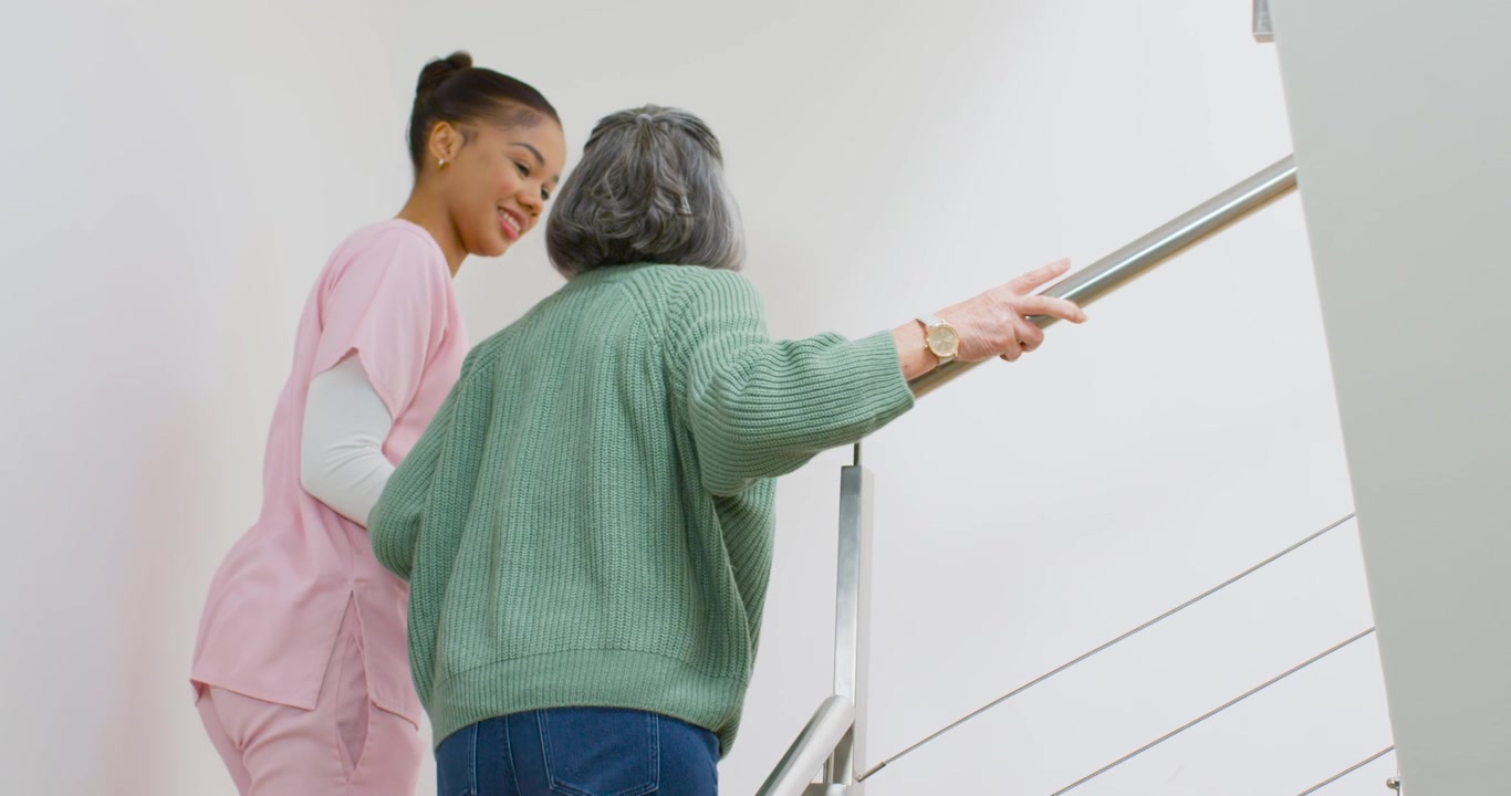 Diverse female carer in pink scrubs aiding senior in green sweater climbing steps and holding rail