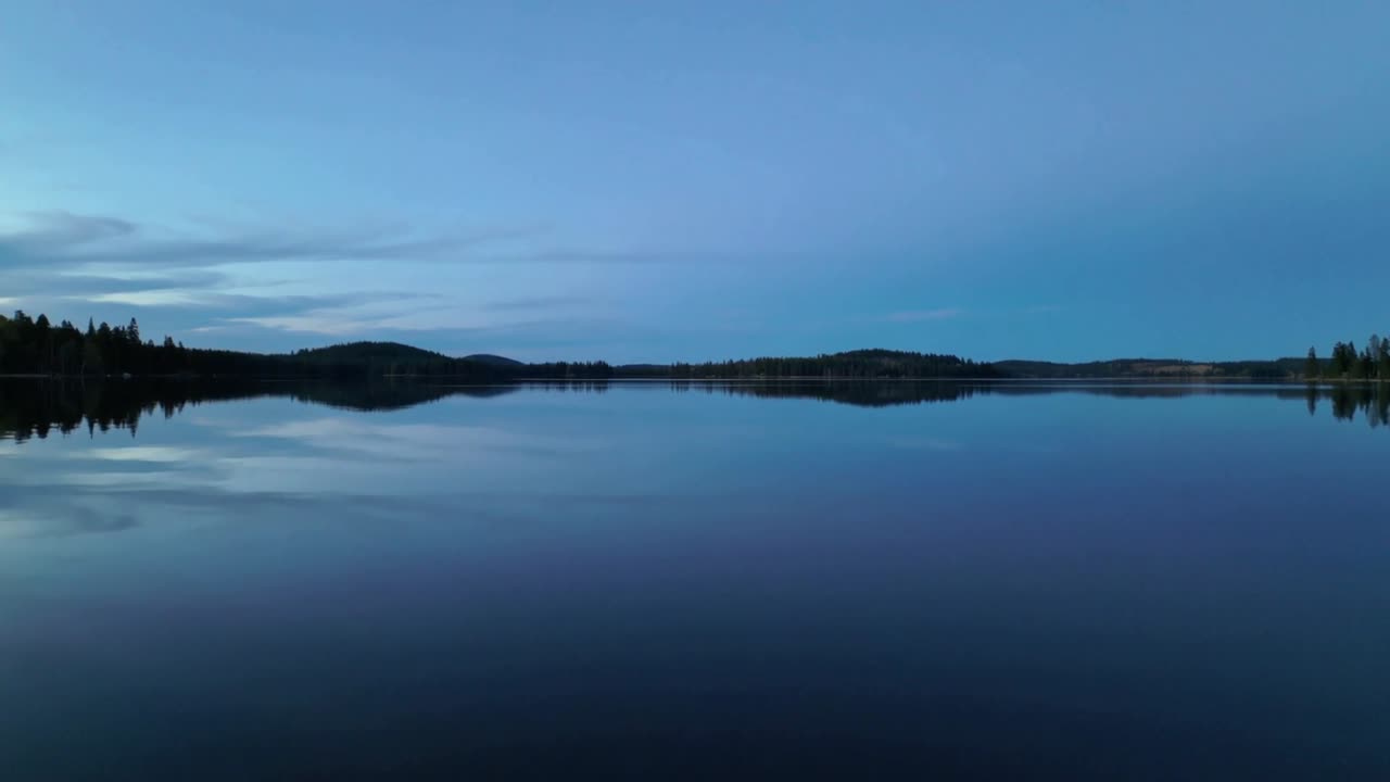 Reflecting lake surface mirroring pale blue sky and treeline at rural shore, dawn light rising