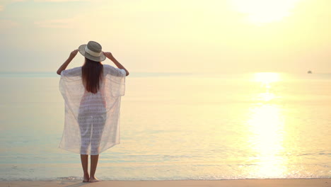 Mujer-Asiática-Solitaria-En-Túnica-Transparente-De-Verano-Y-Sombrero-En-La-Playa-De-Arena-En-El-Sol-Dorado-De-Verano-Sobre-El-Mar-Tropical,-Fotograma-Completo-Estático