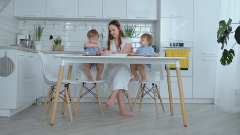 Young-beautiful-mom-in-a-white-dress-with-two-children-are-smiling-and-eating-fresh-burgers-in-their-kitchen