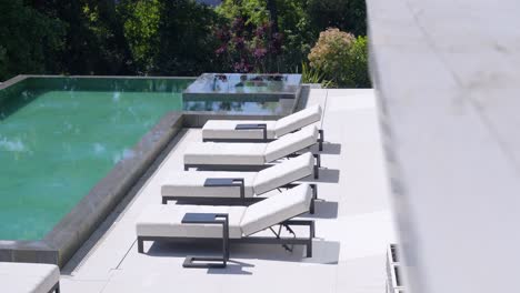 view from the balcony of a modern and luxurious white and gray terrace with an infinity pool, neatly lined sun loungers and nature in the background