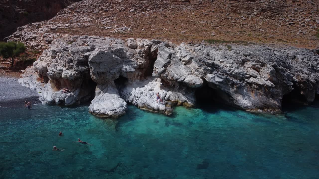 People Swimming In Turqoise Waters Exploring Caves In Rocks In Marmara beach, Crete Greece
