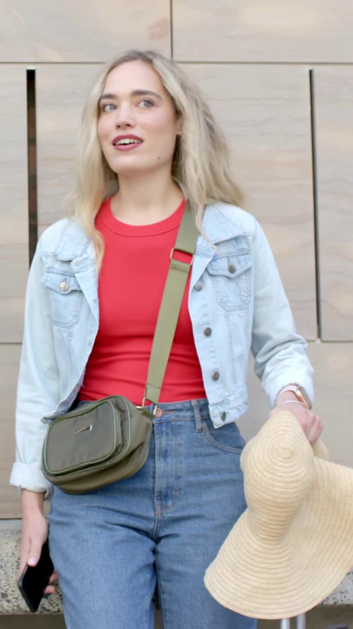 Vídeo vertical: Mulher entrando no quadro, segurando chapéu de palha e telefone, vestindo jeans, reagindo fora da tela.