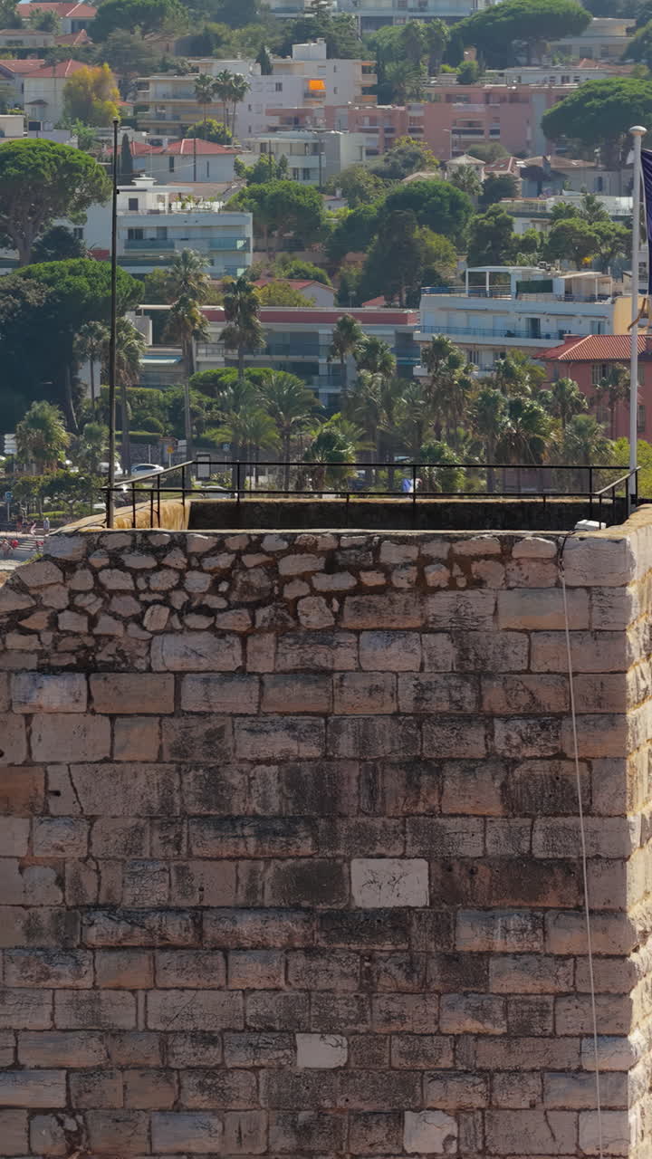 Aerial drone view of the French flag waving above Antibes Old Town medieval tower, with palm-lined promenade and the Mediterranean Sea in the background. Vertical