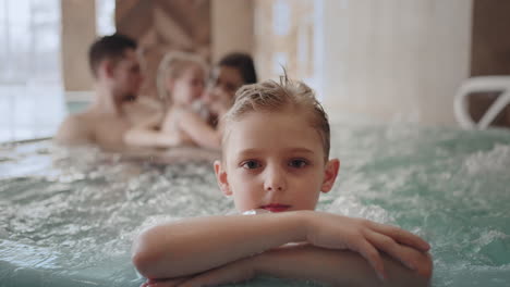 Un-Niño-Lindo-Está-Mirando-La-Cámara-En-Un-Parque-Acuático,-La-Familia-Está-Tomando-Un-Baño-Caliente-Y-Escalofriante-En-La-Piscina