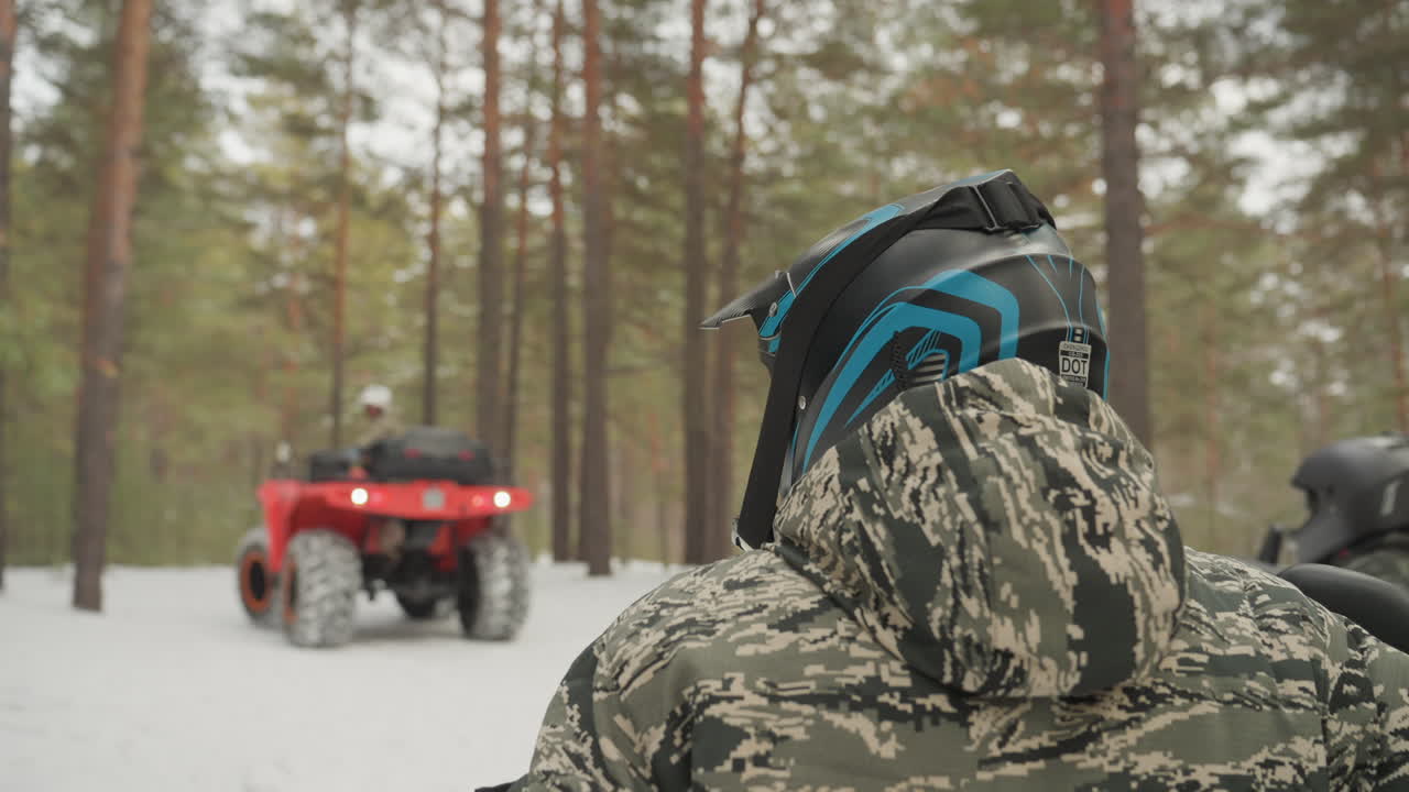 Un pilota di ATV in una pineta innevata osserva un quad rosso avvicinarsi. Primo piano posteriore del pilota con giacca mimetica e casco blu, sotto una luce invernale soffusa, tra pini alti e morbide tracce sulla neve. Si percepisce il rombo del motore e una tesa anticipazione.