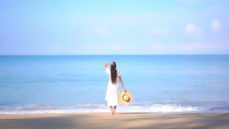 Mujer-Asiática-Parada-En-La-Playa-Junto-Al-Mar-Y-Levantando-Las-Manos-Usando-Un-Vestido-Blanco-De-Verano-Y-Sosteniendo-Un-Sombrero-De-Paja-En-La-Mano---Espacio-De-Copia-De-La-Vista-Posterior-De-La-Plantilla-De-Vacaciones