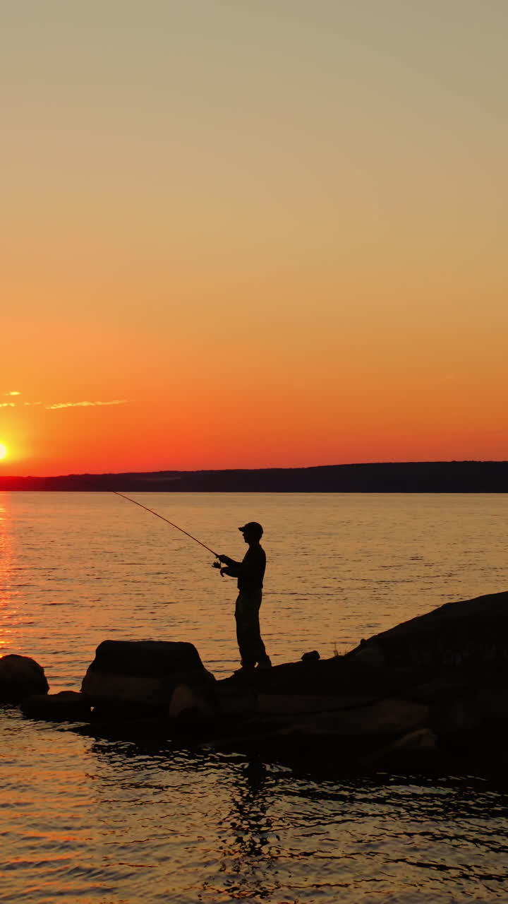 Man fishing at sunset. Silhouette of a man with fishing rod standing on the stone in water in the evening. Fishermen on the lake against orange sunset. Vertical video