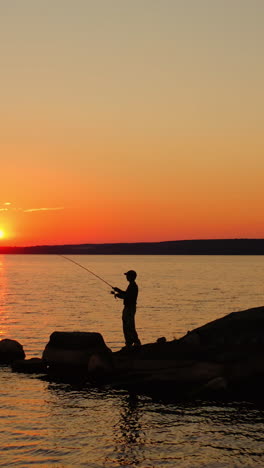 man fishing at sunset. silhouette of a man with fishing rod standing on the stone in water in the evening. fishermen on the lake against orange sunset. vertical video