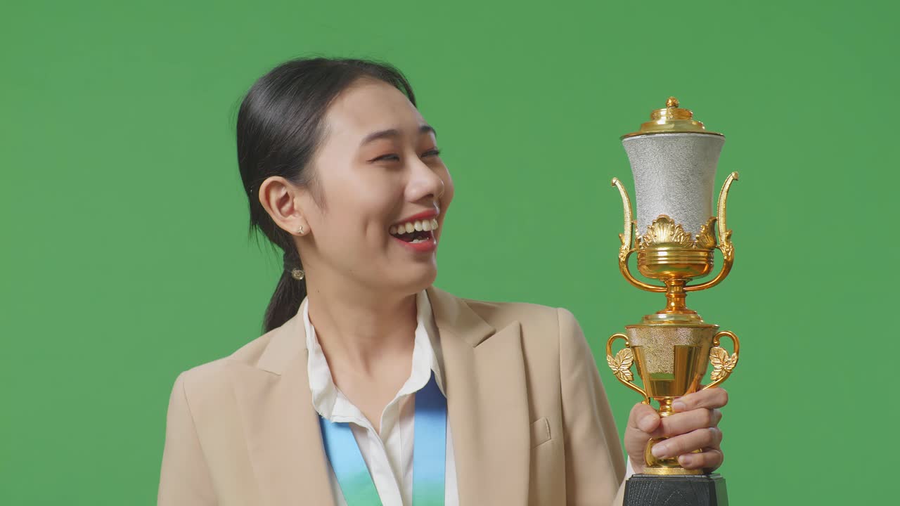 슈트를 입은 아시아 사업가 여성은 금메달을 입고 손에 금 트로피를 들고 첫 번째 우승자로 스튜디오의 녹색 화면 배경에서 카메라에 미소 짓고 있습니다.