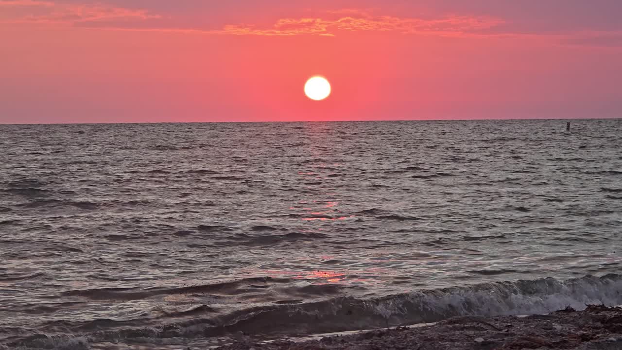 The sun sets low over tranquil sea waves, casting warm pink and orange tones across the sky and reflecting shimmering light on the water near the shore.
