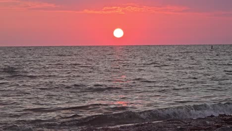 the sun sets low over tranquil sea waves, casting warm pink and orange tones across the sky and reflecting shimmering light on the water near the shore.