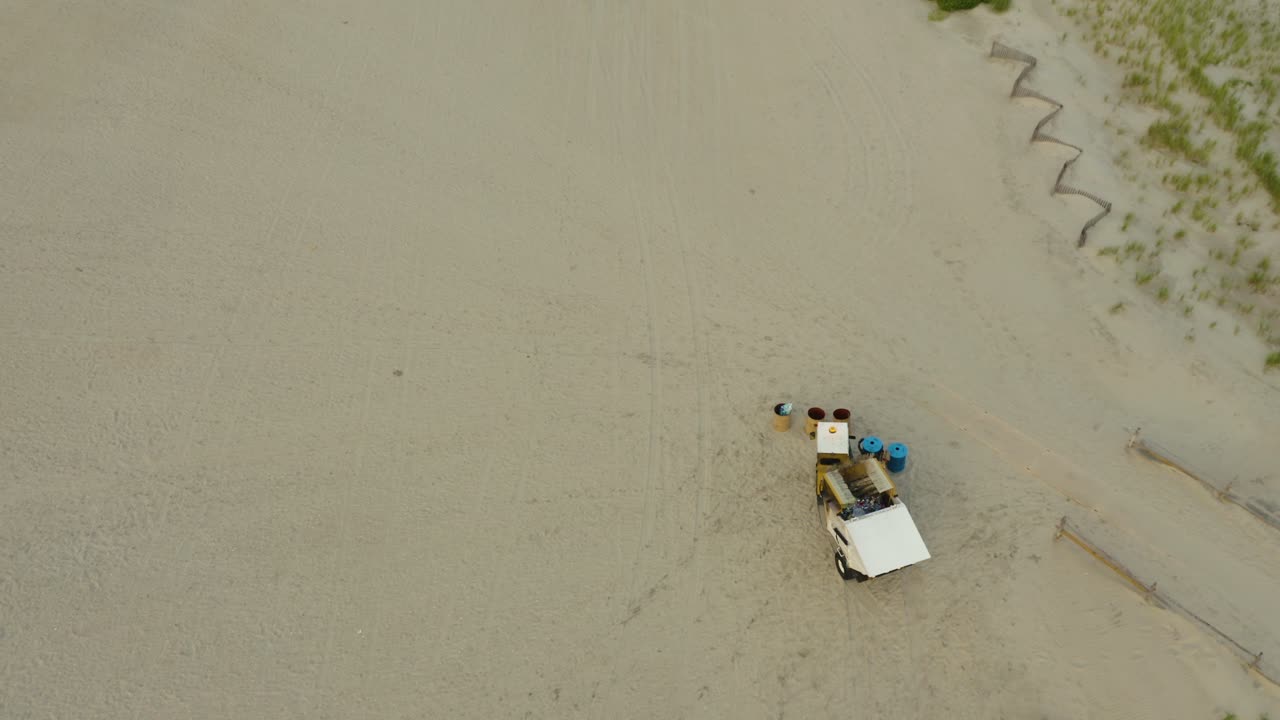 Drone shot, garbage collection buggy emptys bins on beach then drives off into distance