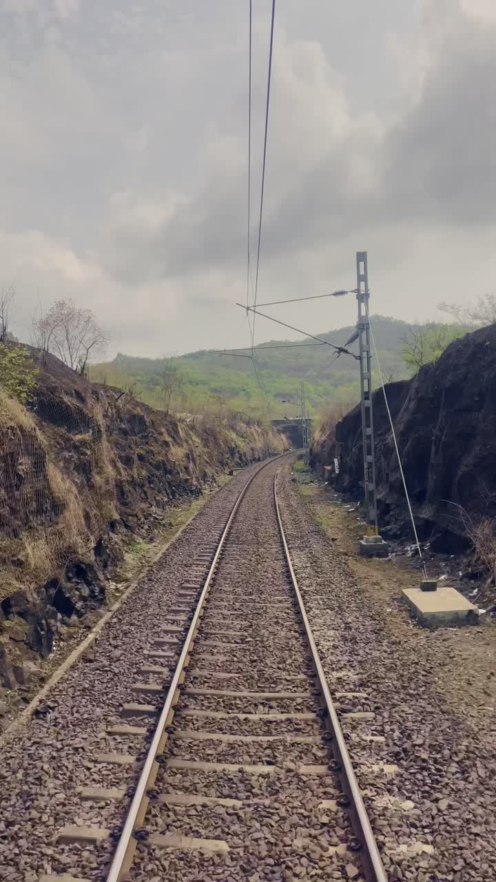 Train tracks stretch forward, flanked by gentle, grassy hills on both sides. The sky above is bright yet scattered clouds Maharashtra India 4K