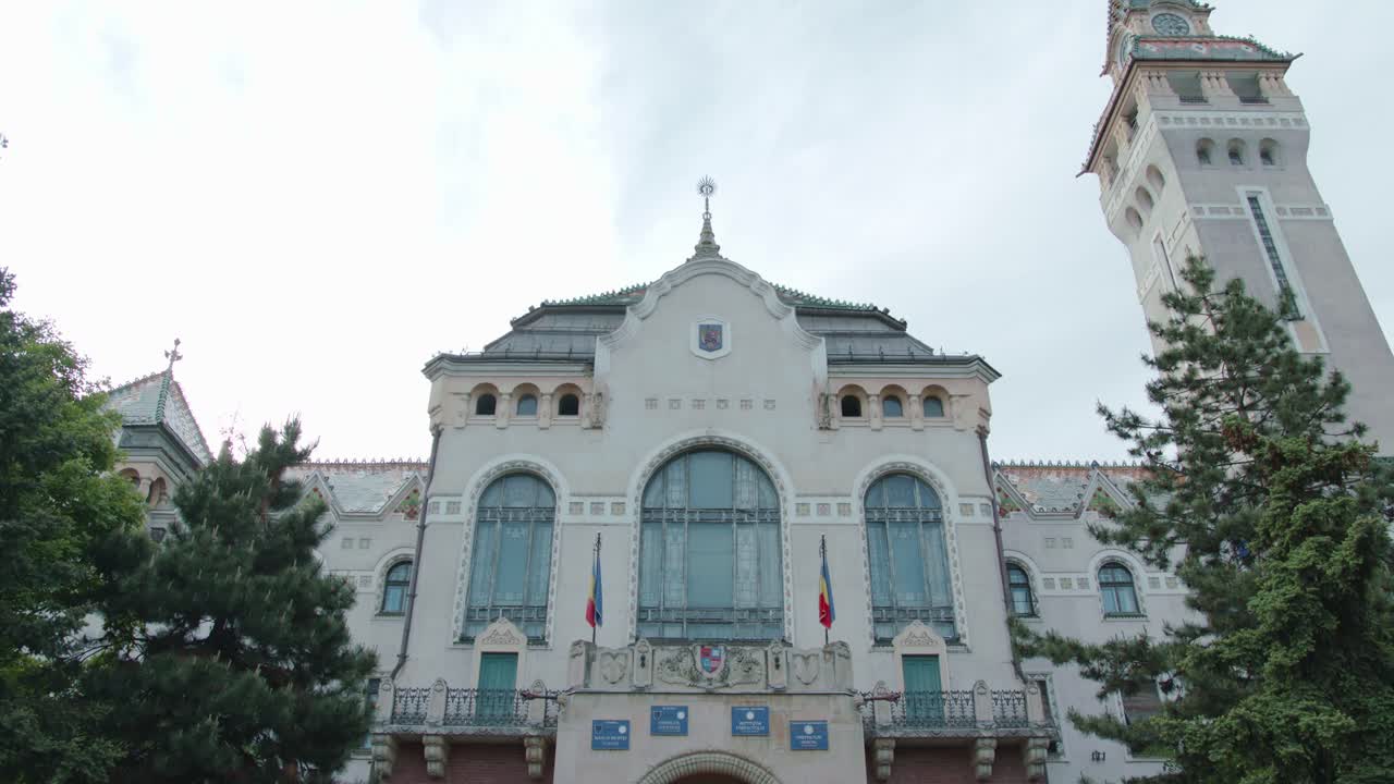 A slow, majestic tilt-up shot of the historic Palace of Culture in Targu Mures. The camera reveals the grand Art Nouveau facade, entrance, and iconic clock tower