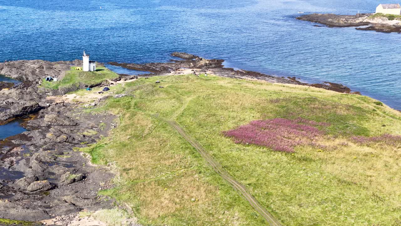 Drone glides toward white lighthouse, rocky shoreline, wildflowers, and blue sea in bright daylight