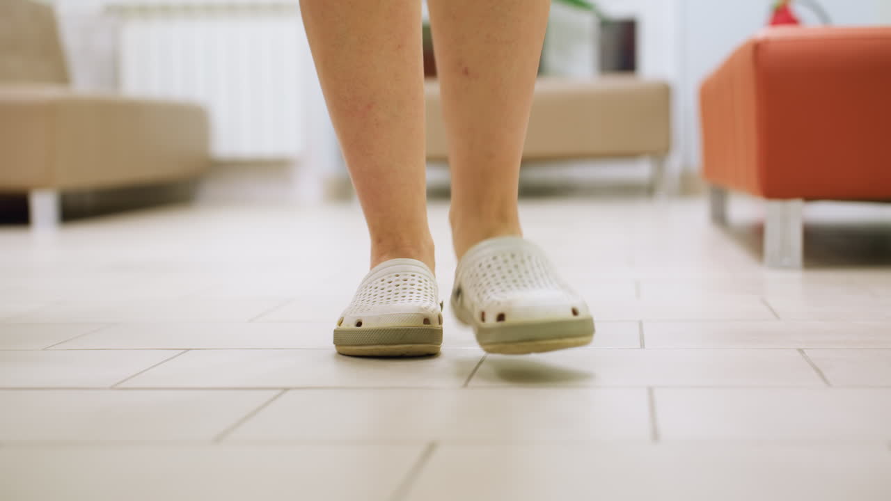 Rear leg view of woman in white crocs walking across tiled floor through reception area with couches visible in background clean interior setting suggests clinical or institutional environment