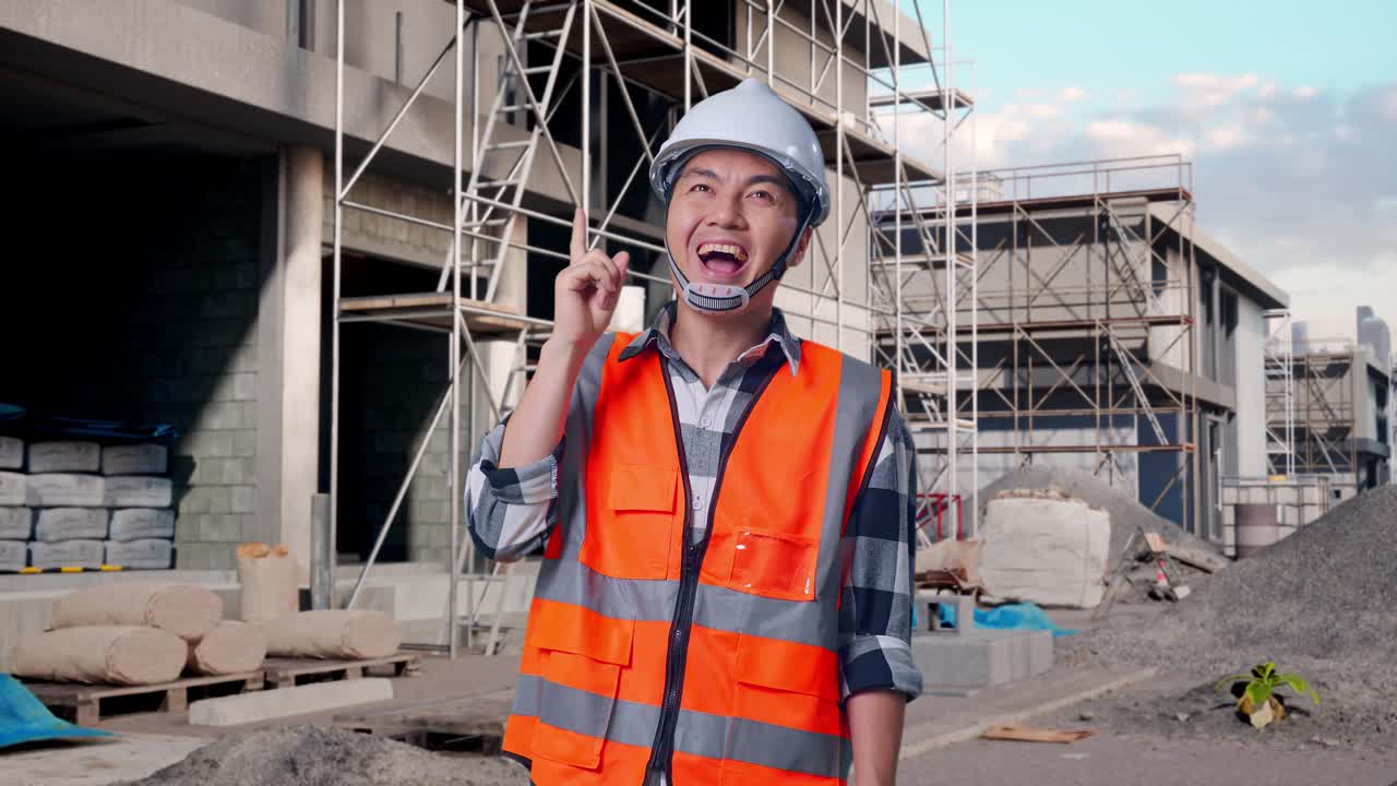 Asian Male Engineer With Safety Helmet Thinking And Looking Around Then Raising His Index Finger While Standing At Building & Construction Site in Progress to New House