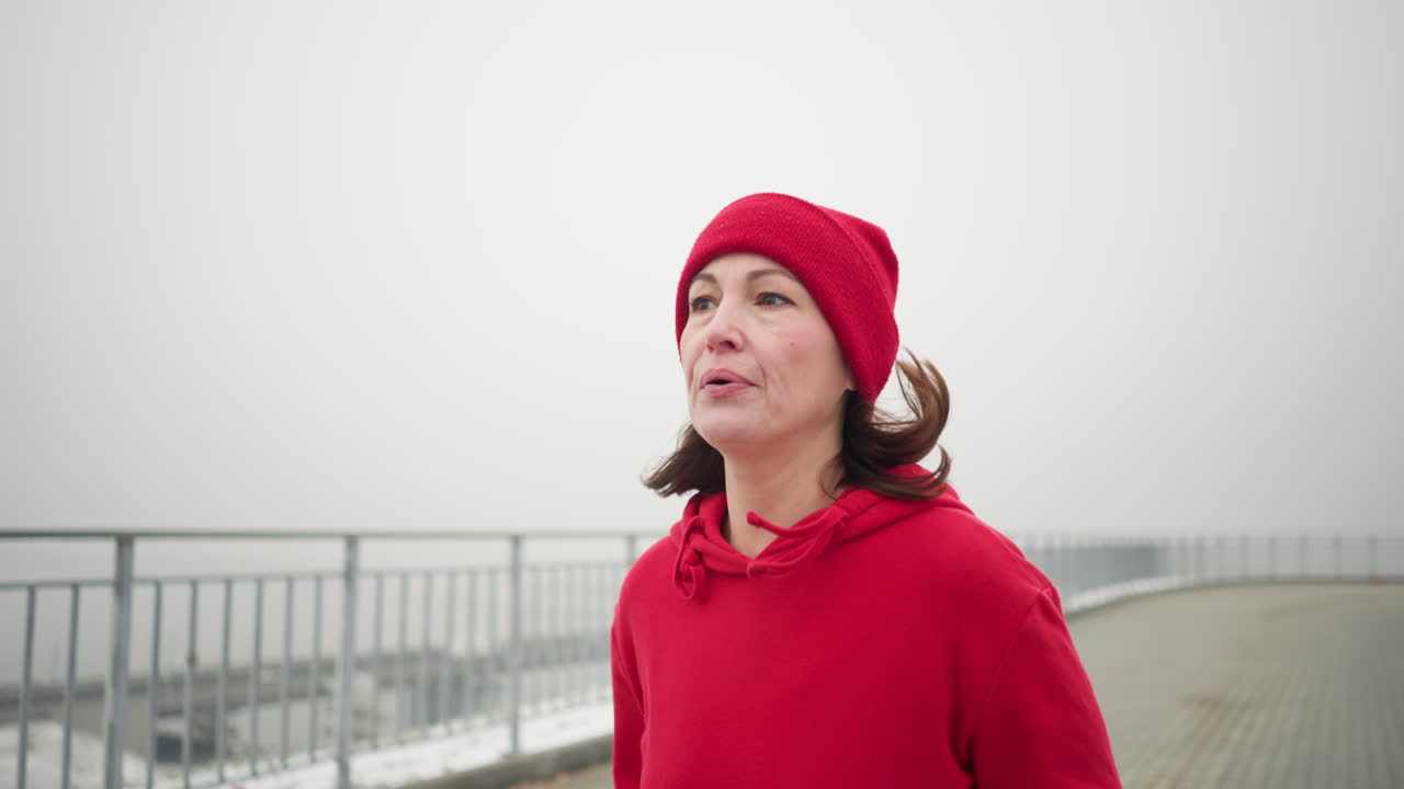 madre cansada con gorra roja y sudadera con capucha corriendo por el camino cerca de la barandilla de hierro, puente lejano con coches visibles, destacando el estilo de vida activo, el fitness de invierno y el paisaje urbano con una atmósfera de niebla