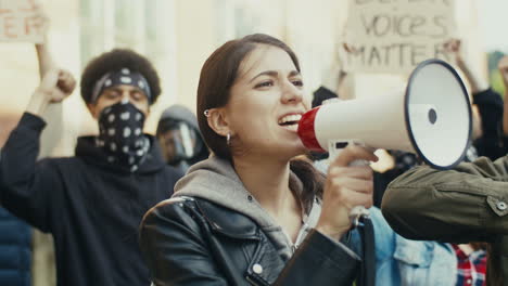 mujer caucásica hablando por un altavoz en una protesta con un grupo multiétnico de personas en la calle