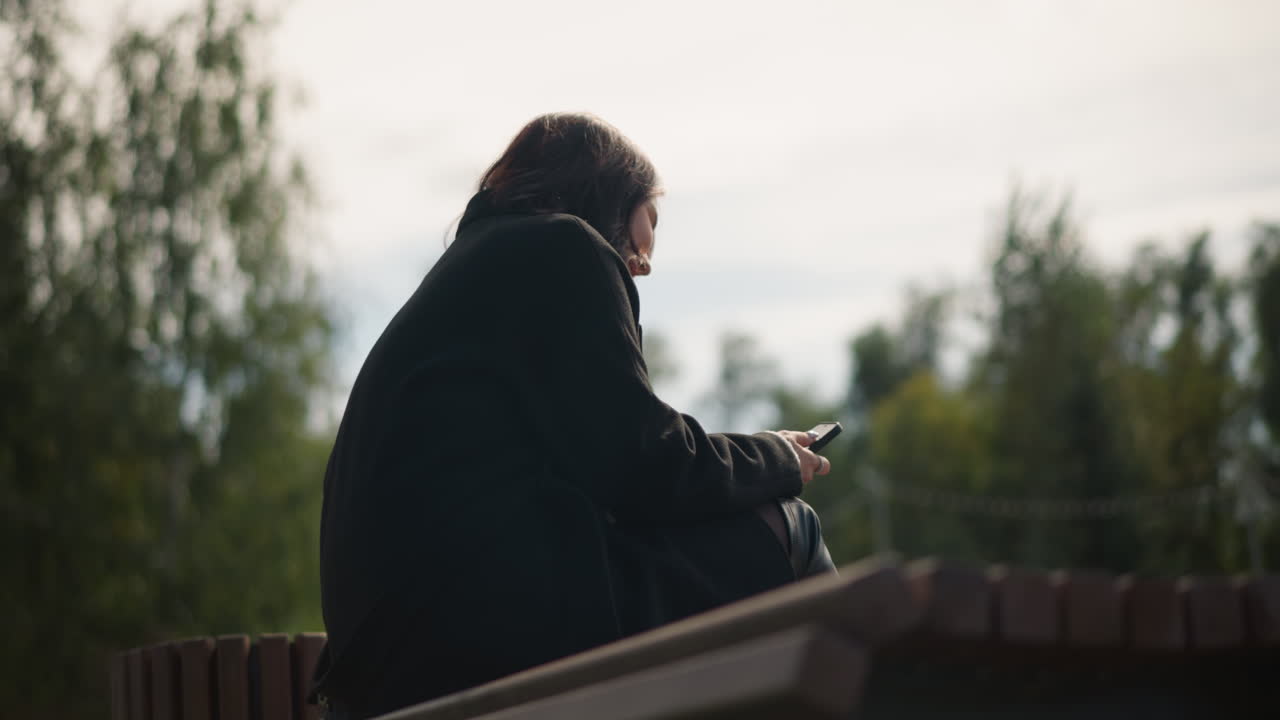 Back view of woman in black coat smiling while using smartphone outdoors, seated against blurred natural background with trees and soft sunlight