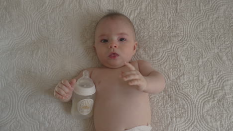 newborn baby with his milk bottle gesturing, laying on back on white bed sheets