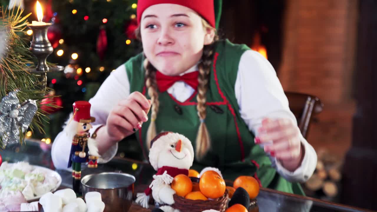 Joyful girl elf in green costume eating at table. Greedy female elf taking all sweets from plate for herself