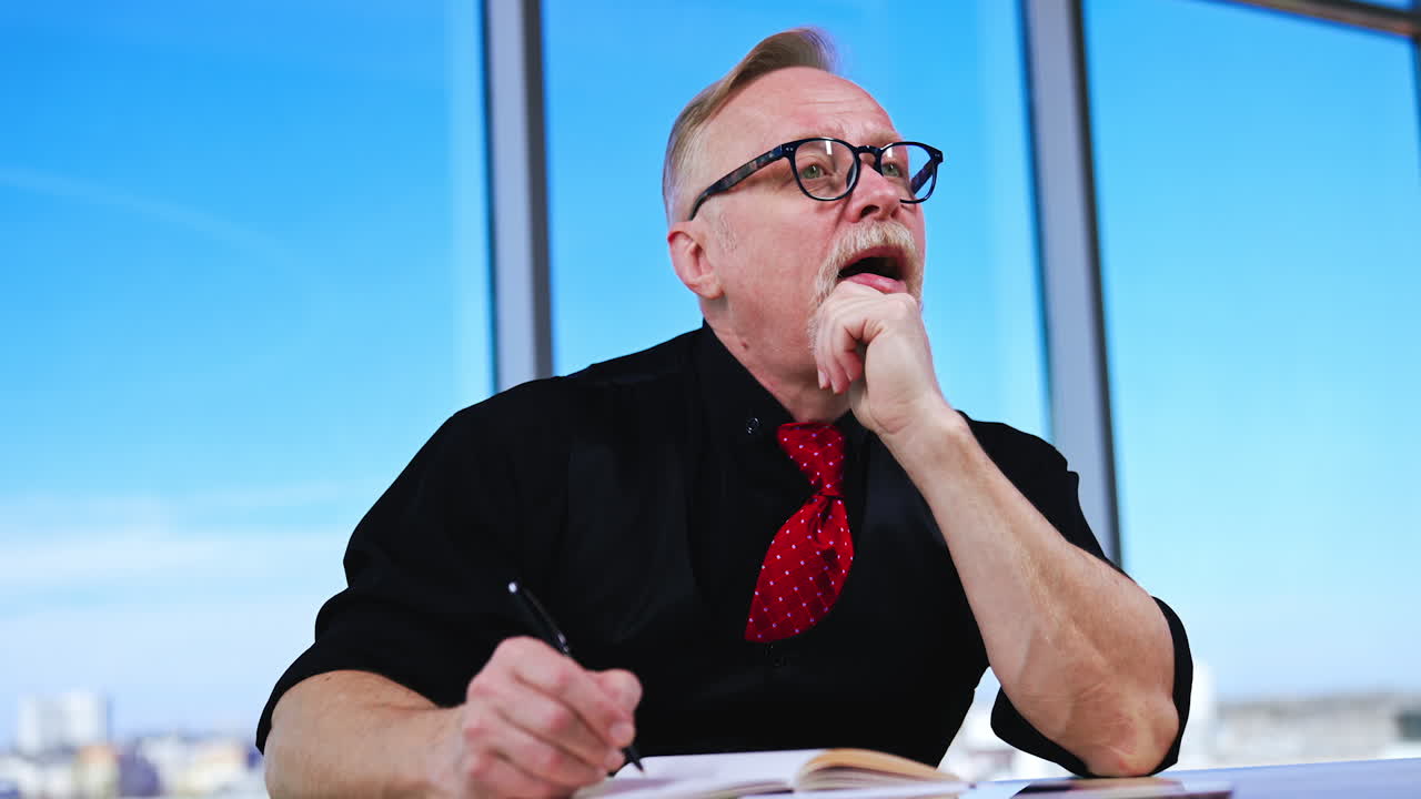 Man wearing black shirt, vest and red tie stops writing. Man thinks for a while, crosses written and continues to write.