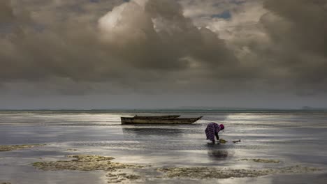 Mujer-De-Zanzíbar-Recogiendo-Mariscos-Del-Mar-Durante-La-Marea-Baja-Con-Un-Barco-De-Pesca-En-Segundo-Plano.