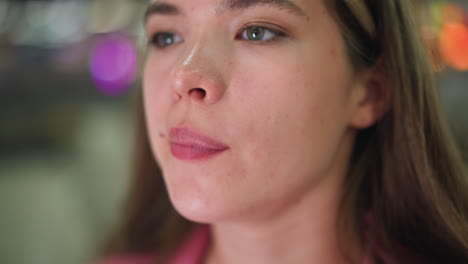 primer plano de una mujer lamiendo sus labios rosados como si tuviera algo en la boca se ven manchas en su cara, con luces borrosas de colores parpadeando en el fondo, y un objeto poco claro detrás de ella