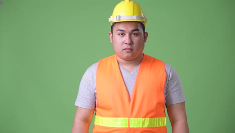 young handsome overweight asian man construction worker against green background