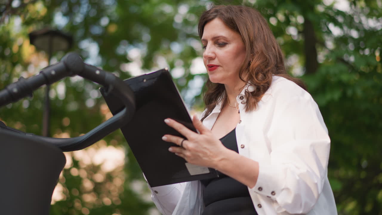 Caucasian woman reading decree outdoors, holding tablet and speaking into microphone at park podium, formal white jacket, focused delivery, evening light and greenery, community leader presenting