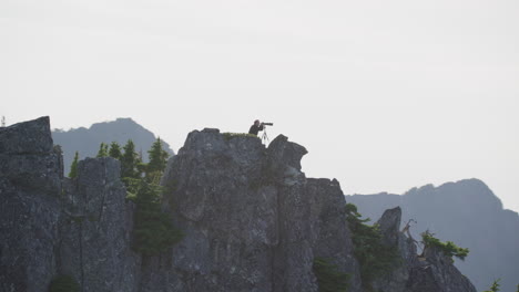 photographer kneels on mountain ridge as silhouette against hazy sky, tim durkan wide tracking helicopter counterclockwise orbit slow motion