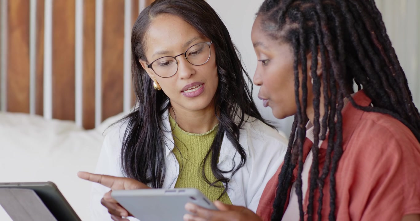 Two female colleagues leaning in, right woman gesturing and explaining tablets on bed, reviewing