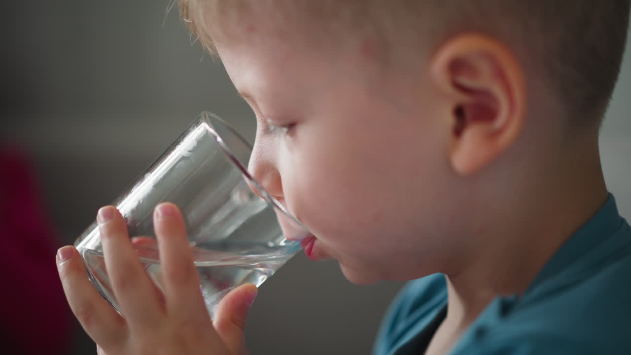 Close up side view of cute young boy wearing blue shirt drinking water from clear glass indoors with partial view of person in red top in background