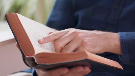 closeup view of male hands holding book. young man is reading a book while sitting in white chair, at home. window and green plants on background