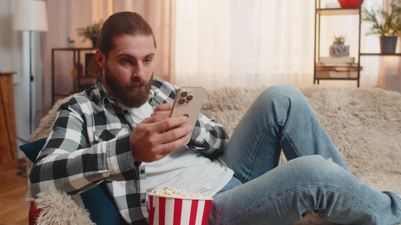 Young man on sofa with popcorn smartphone scroll sudden wow reaction shock moment home surprise