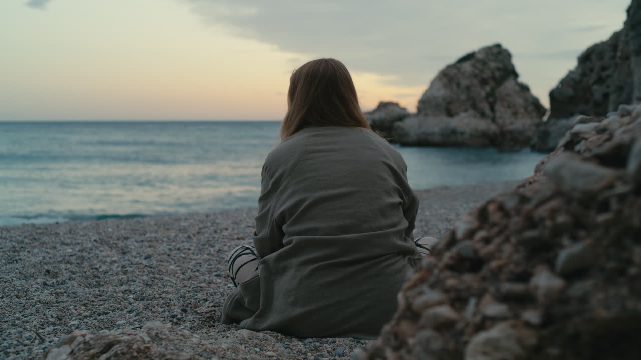 Vista da dietro di una giovane donna seduta su una tranquilla spiaggia di ciottoli al tramonto, che guarda verso il mare calmo e la costa rocciosa nella tenue luce dell'ora blu.
