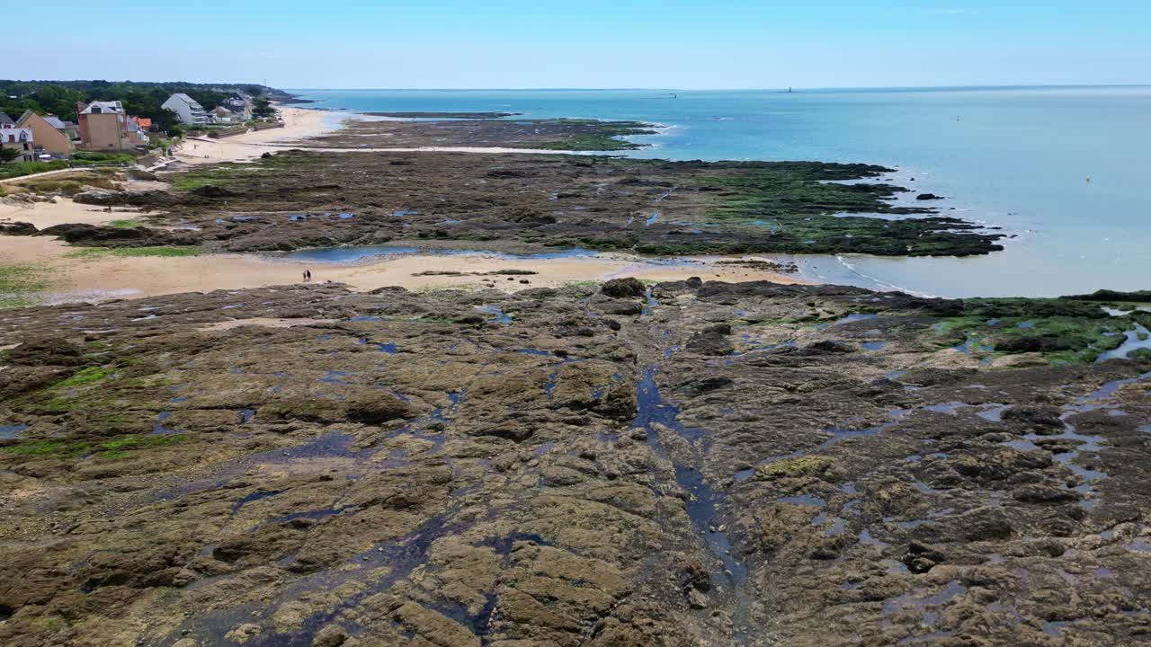 Forward aerial drone view of Bonne Source Beach in Pornichet, flying above rocky shores with the sea on the right and houses with vegetation on the left - Brittany France