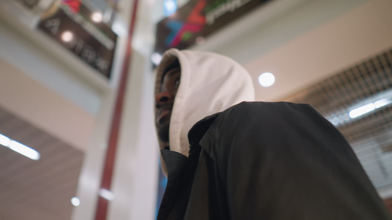 Young man in hoodie looking up in modern shopping mall with soft lighting, wearing shoulder bag, gazing at ceiling and advertisements, abstract and calm atmosphere, commercial building interior