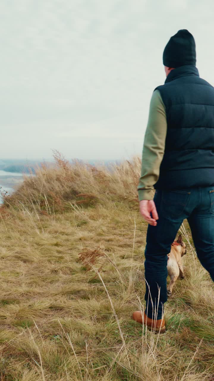 En person, der nyder en fredfyldt gåtur med sin hund i et græsrigt landskab, hvilket viser båndet mellem menneske og kæledyr i en rolig, naturlig omgivelse midt i smuk natur