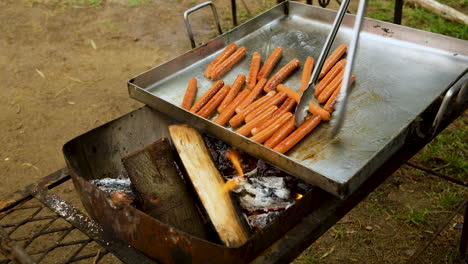 Freír-Salchichas-En-Una-Gran-Sartén-Para-Acampar-En-Un-Fuego-De-Leña-Abierto-Usando-Pinzas