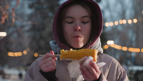 lady enjoying maize held by stick wrapped in white sachet with bokeh lights in blurred background, steam rising from fresh corn with people walking in background