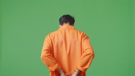 prisionero masculino asiático con esposas de pie en el fondo de la pantalla verde, girándose para mirar a la cámara enojado y malhumorado